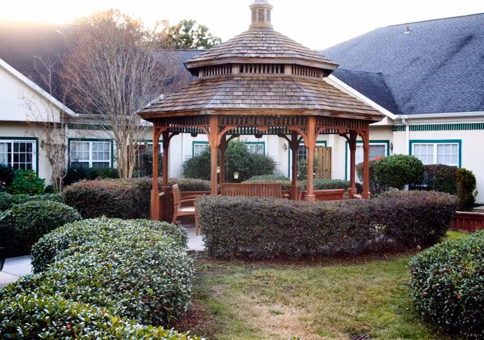A wooden gazebo with a shingled roof surrounded by neatly trimmed bushes and greenery in the courtyard of a senior living facility, with building windows and doors visible in the background.