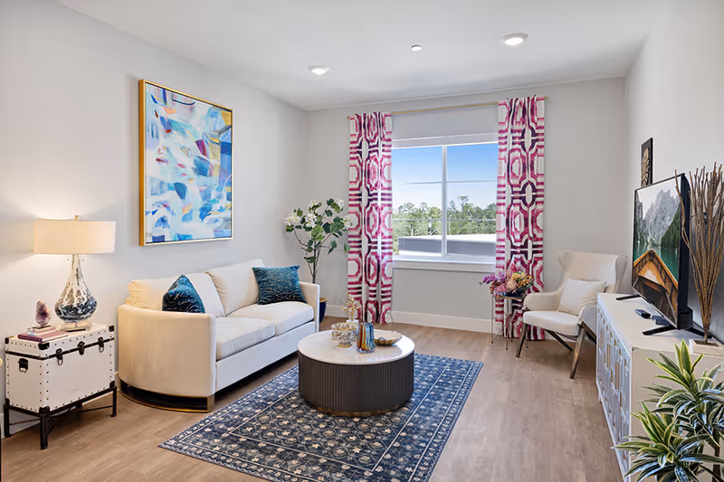 A bright and modern living room with a white sofa adorned with blue pillows, a round coffee table on a patterned blue rug, a white armchair, a large window with pink and white patterned curtains, a TV on a white cabinet, a table lamp on a white trunk, and decorative plants and artwork on the walls.