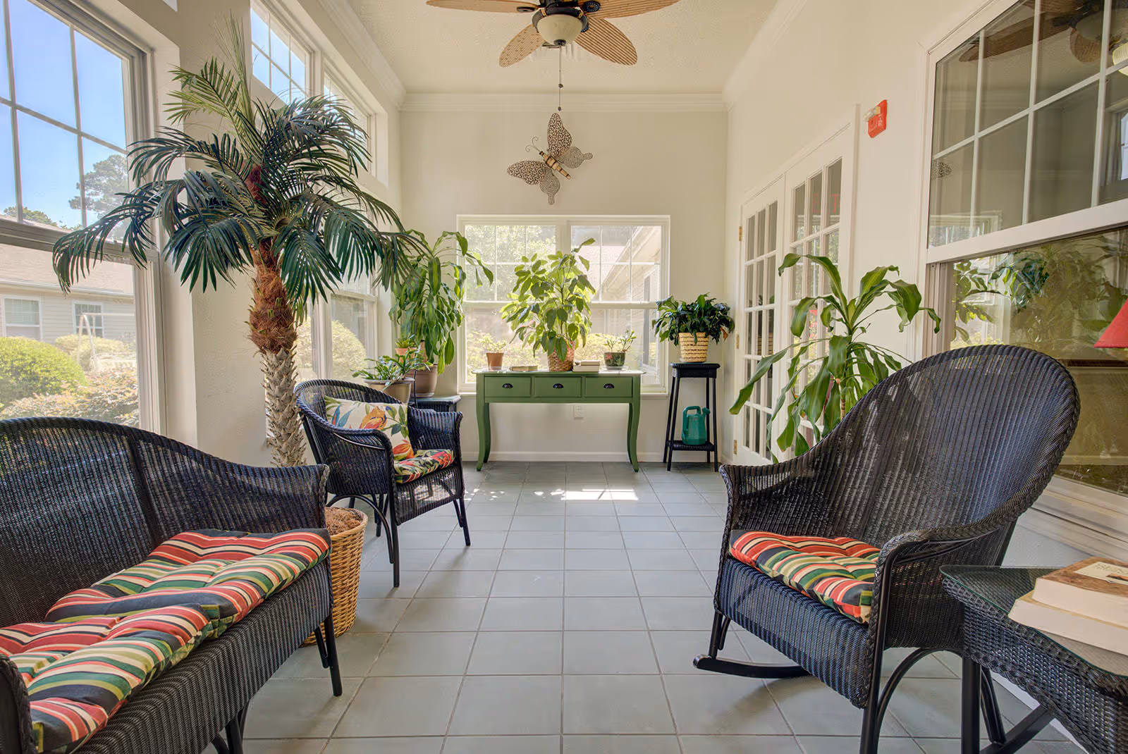 A bright sunroom with large windows letting in natural light, featuring black wicker furniture with colorful striped cushions, several potted plants, a green table with drawers, and a ceiling fan with a light. The room has tiled flooring and white walls, creating a cozy and inviting sitting area.
