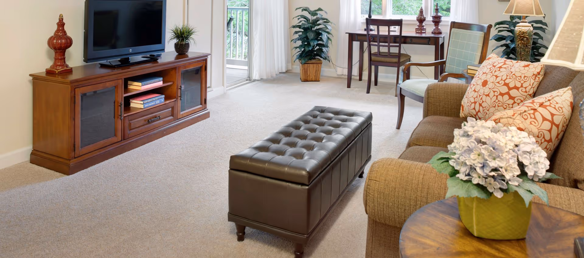Bright furnished living room with a TV on a wooden console, a tufted leather ottoman, a sofa with patterned pillows, a side table with flowers, and a small desk by the window.