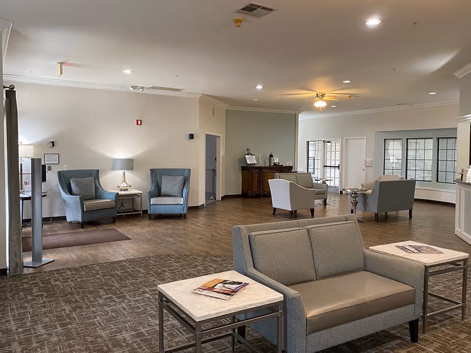 A spacious senior living facility common area with multiple seating arrangements including cushioned armchairs and sofas. The room features wood and carpet flooring, soft lighting from lamps and ceiling lights, and a sideboard with coffee supplies in the background. Large windows with blinds allow natural light into the space.