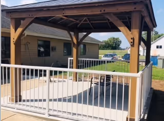 A wooden gazebo structure with a peaked roof situated on a concrete patio area surrounded by a white metal railing. In the background, there is a single-story building, parked cars, and a grassy area under a blue sky with some clouds.