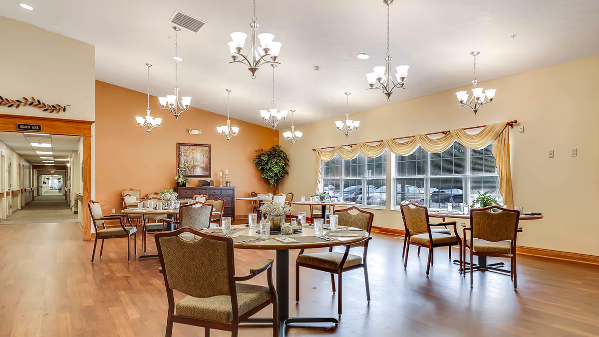 A bright and spacious dining room in an assisted living facility with multiple round tables set with placemats, glasses, and silverware. The room features wooden flooring, beige and orange walls, large windows with draped curtains, several chandeliers hanging from the ceiling, and a hallway labeled 'Hidden Cove' visible in the background.