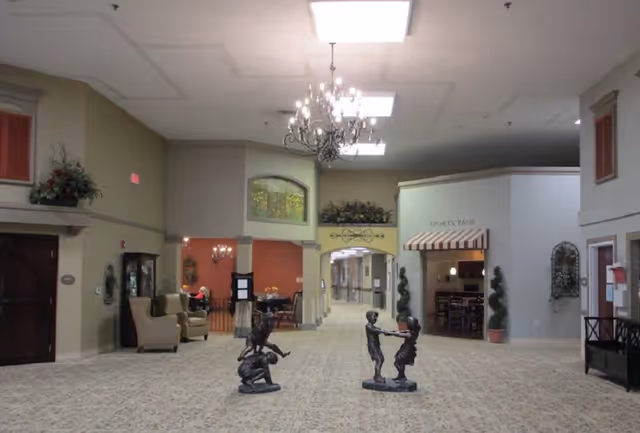 Open senior living facility lobby with chandeliers, seating areas, decorative sculptures, and a central corridor to activity rooms.