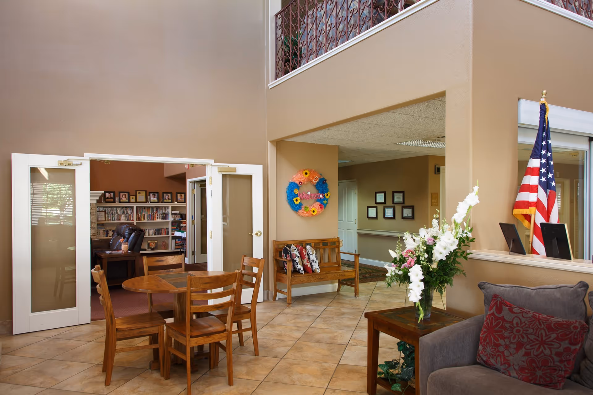 Interior view of a senior living facility common area with a wooden table and chairs in the foreground, a wooden bench with colorful pillows against a wall with a decorative welcome wreath, an American flag near a windowed reception area, and a cozy seating area with a gray armchair and floral cushions. In the background, there is a room with bookshelves and framed pictures on the wall.