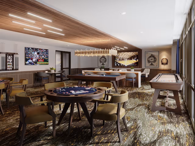 A spacious senior living facility game room with multiple gaming tables including a poker table, pool table, and shuffleboard. The room features comfortable chairs, a dartboard on the wall, two large flat-screen TVs displaying sports, and a modern wooden ceiling design with recessed lighting. Large windows on one side allow natural light to fill the room.