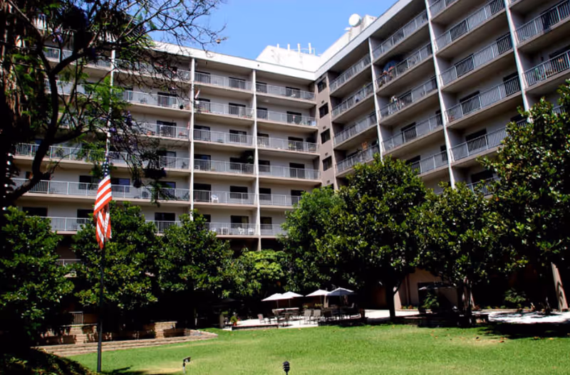 Courtyard with lawn, patio tables and umbrellas surrounded by trees in front of a multi-story residential building with balconies and an American flag.