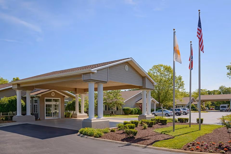 Exterior view of Life Care Center of Sparta showing the main entrance with a covered drop-off area supported by white columns, three flagpoles with flags, landscaped bushes and flowers, and a parking lot with several cars under a clear blue sky.