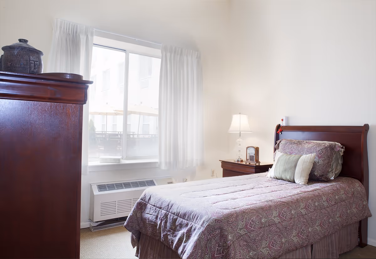 A bright bedroom with a single bed covered in a purple patterned comforter and two pillows. Next to the bed is a wooden nightstand with a lamp and a small clock. A large window with white curtains lets in natural light, and a wooden dresser is partially visible on the left side of the image.