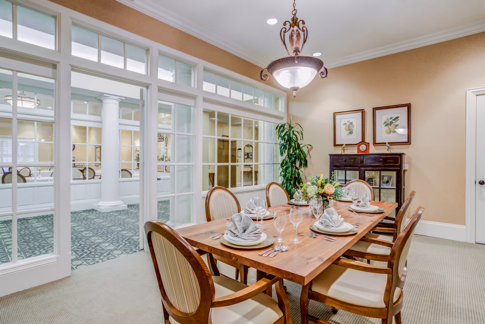 A well-lit dining room with a wooden table set for six people, featuring folded napkins, glassware, and a floral centerpiece. The room has beige walls, a hanging chandelier, framed botanical prints, a dark wooden cabinet, and a large green plant. Glass-paneled double doors open to an adjacent room with patterned carpet and white columns.