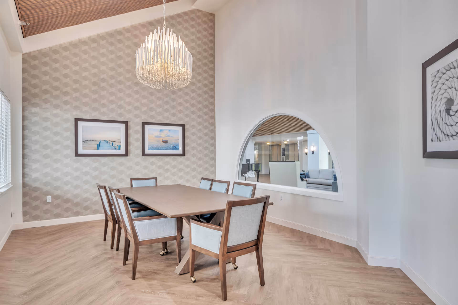 Dining room with a rectangular wooden table surrounded by upholstered chairs, a decorative chandelier, a patterned accent wall, and an arched interior window.
