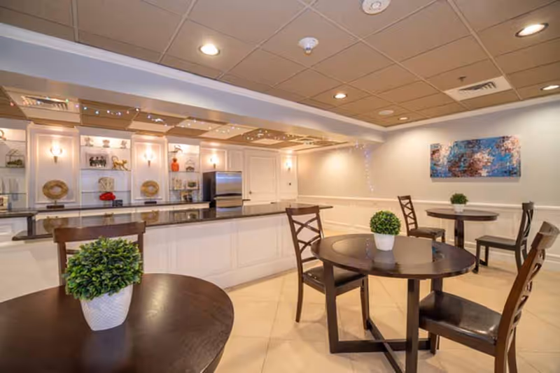 Interior view of a dining area in Tuscany Villa of Naples featuring round wooden tables with small potted plants, wooden chairs, and a kitchen counter with built-in shelves and a refrigerator in the background. The room has a beige tiled floor, a drop ceiling with recessed lighting, and a colorful abstract painting on the wall.