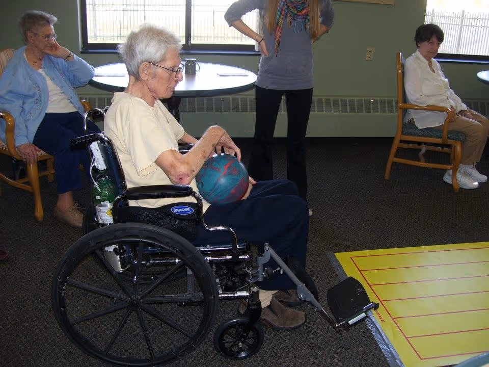 An elderly woman in a wheelchair holding a bowling ball indoors, with two other elderly women seated nearby and a standing person partially visible. The setting appears to be a recreational area in an assisted living facility.
