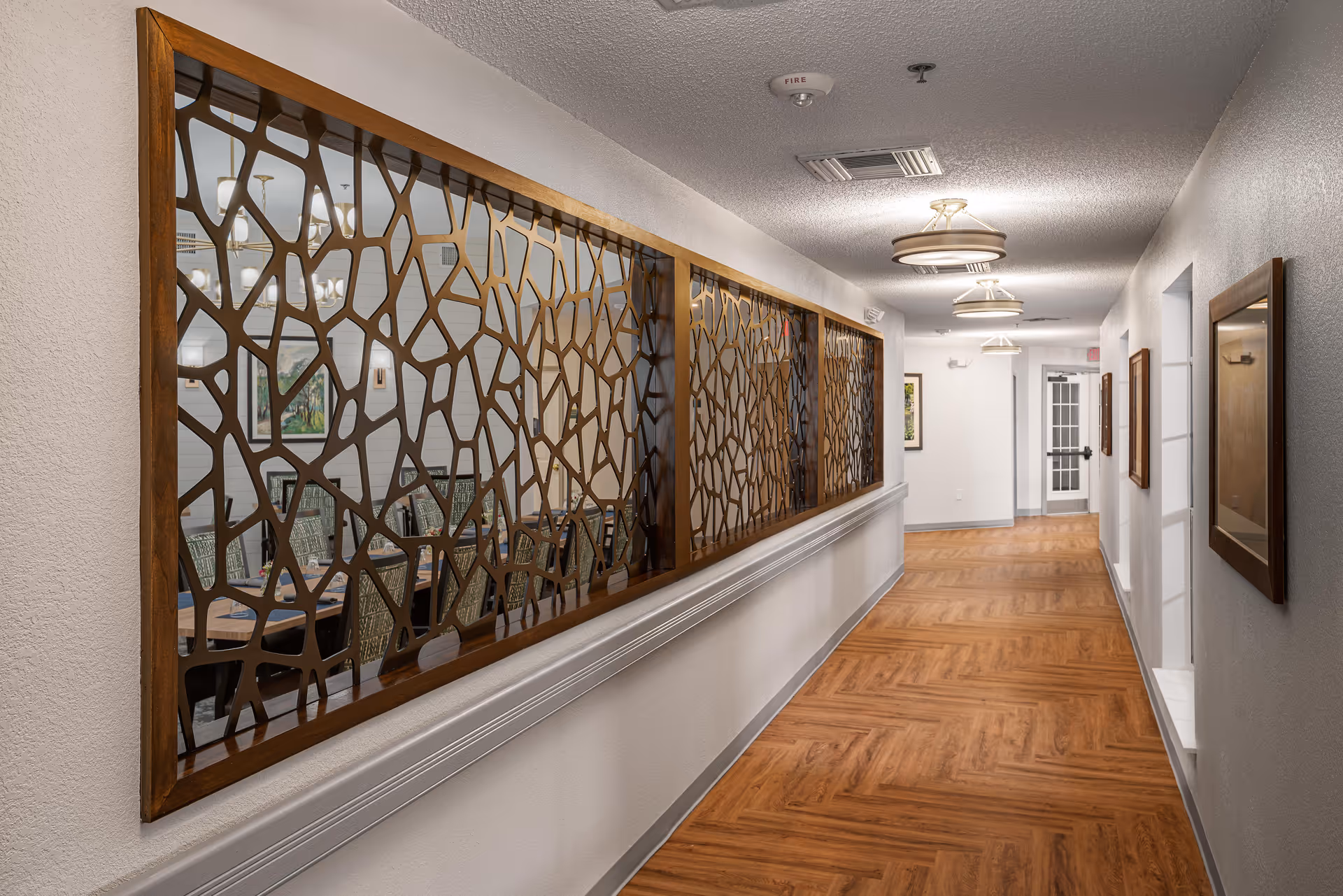 Well-lit corridor in a senior living facility with decorative wooden lattice panels on the left and wood-patterned flooring.