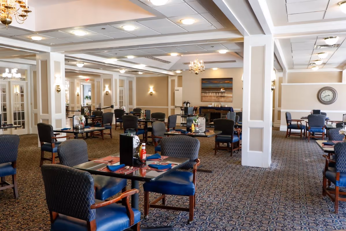 A spacious dining room with multiple tables and chairs arranged neatly. Each table is set with placemats, napkins, and condiments. The room features patterned carpet, beige walls with white trim, chandeliers, and wall sconces providing warm lighting. There is a clock on one wall and a painting on another, with a coffee station visible in the background.