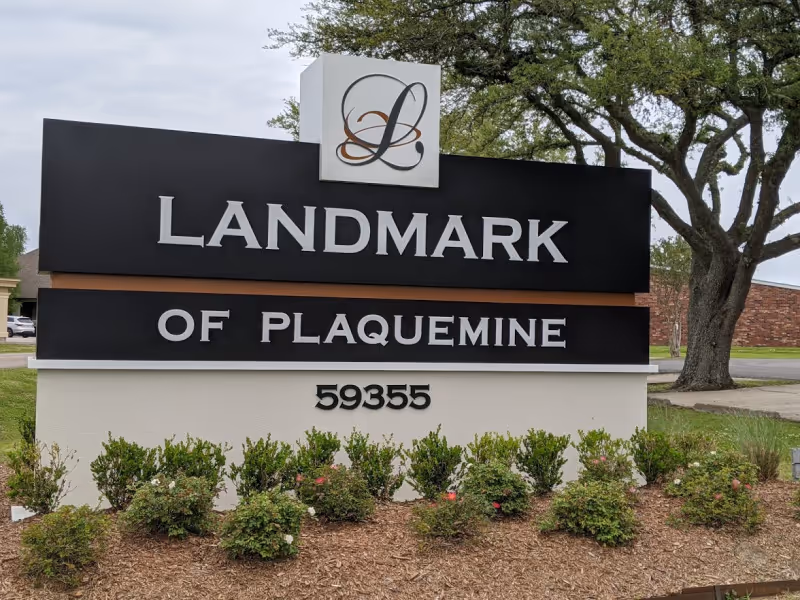 Outdoor sign for Landmark of Plaquemine Nursing Home and Rehabilitation with the address number 59355, surrounded by small bushes and trees in the background.