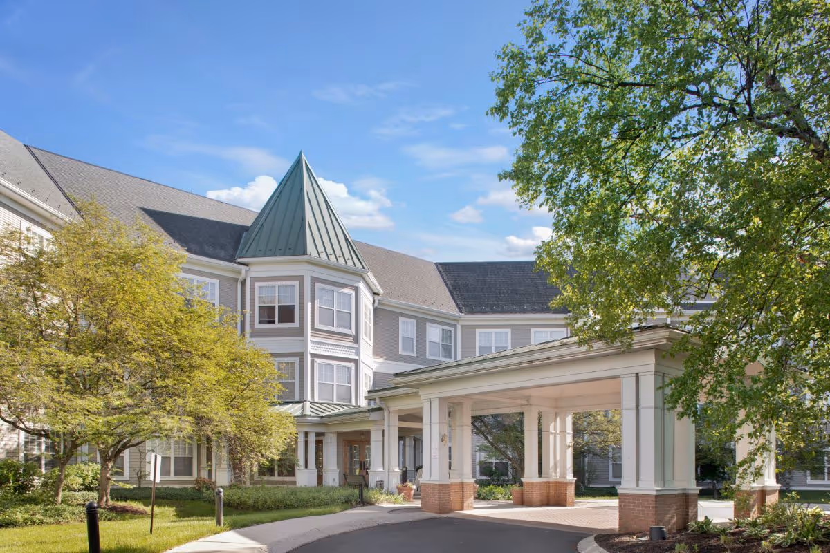 Exterior view of a senior living facility building with a covered entrance, surrounded by green trees and landscaping under a blue sky with some clouds.