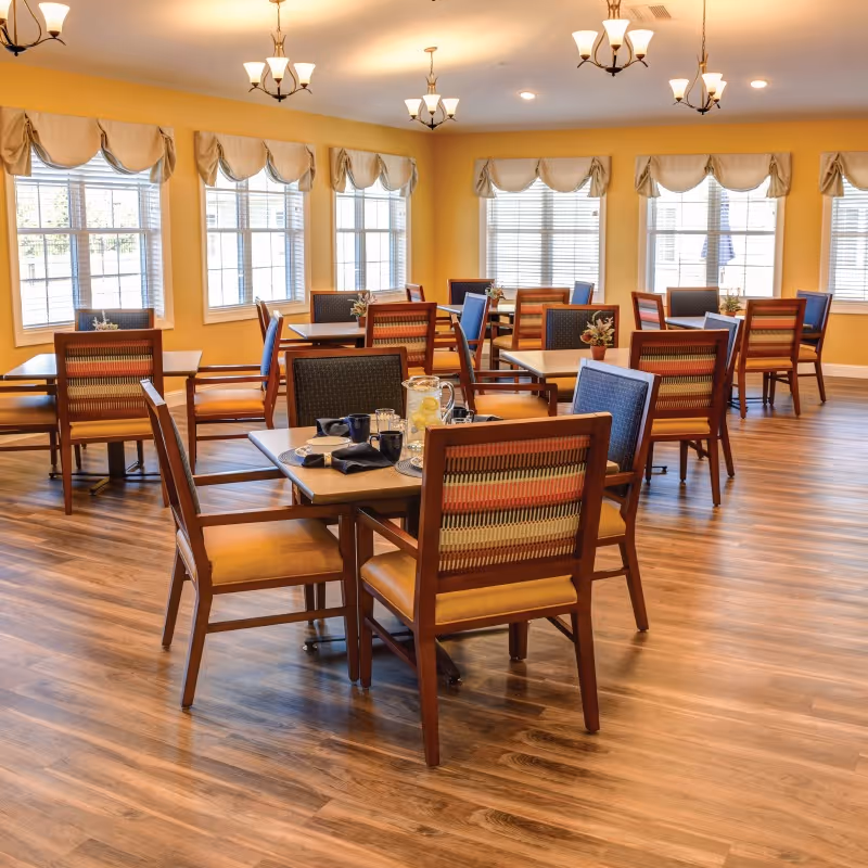A bright dining room with multiple wooden tables and chairs arranged neatly. The room has large windows with white blinds and beige valances, allowing natural light to fill the space. The walls are painted yellow, and the floor has a wood finish. Several chandeliers hang from the ceiling, providing additional lighting. Some tables have pitchers of lemonade and cups set on them.