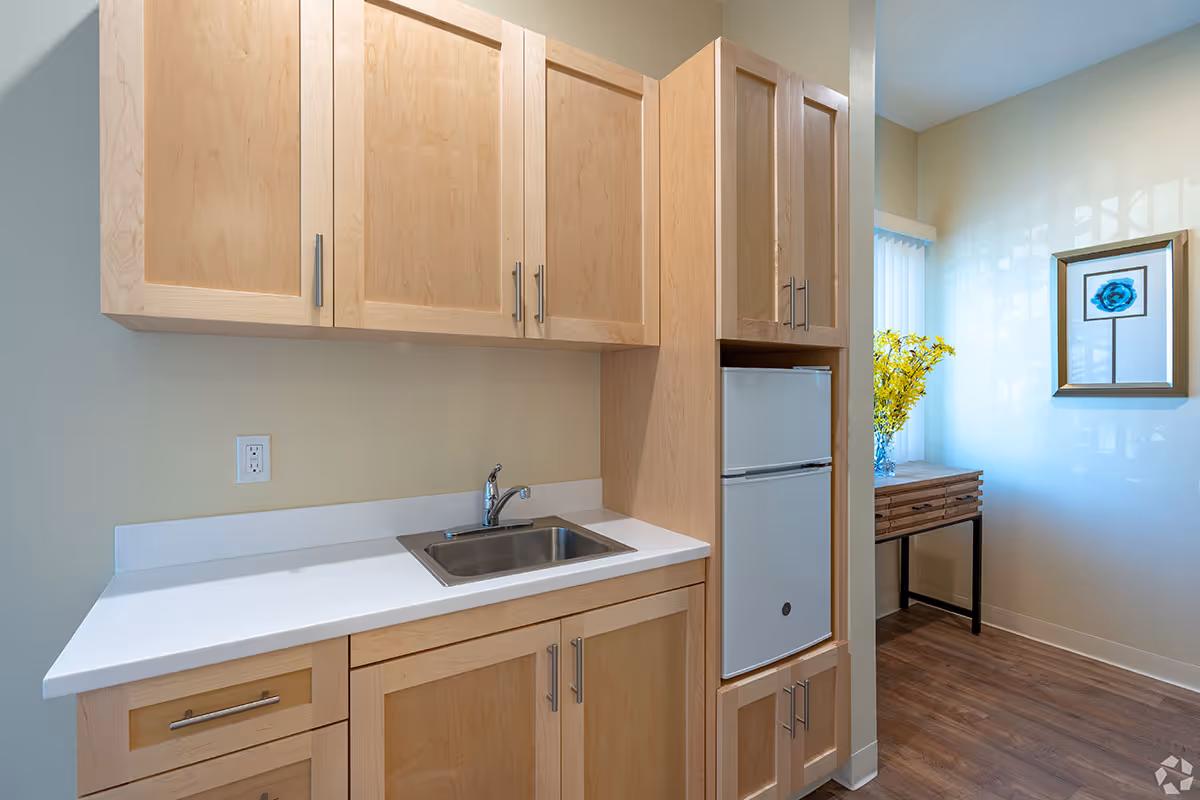 A small kitchen area with light wood cabinets, a white countertop, a stainless steel sink with a faucet, and a compact refrigerator. In the background, there is a wooden table with a vase of yellow flowers and a framed blue flower artwork on the wall. The floor is wood, and the walls are painted light beige.