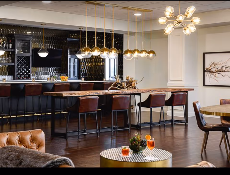 Modern communal lounge and bar area with a long wooden table, leather stools, pendant lighting, and small tables with drinks.