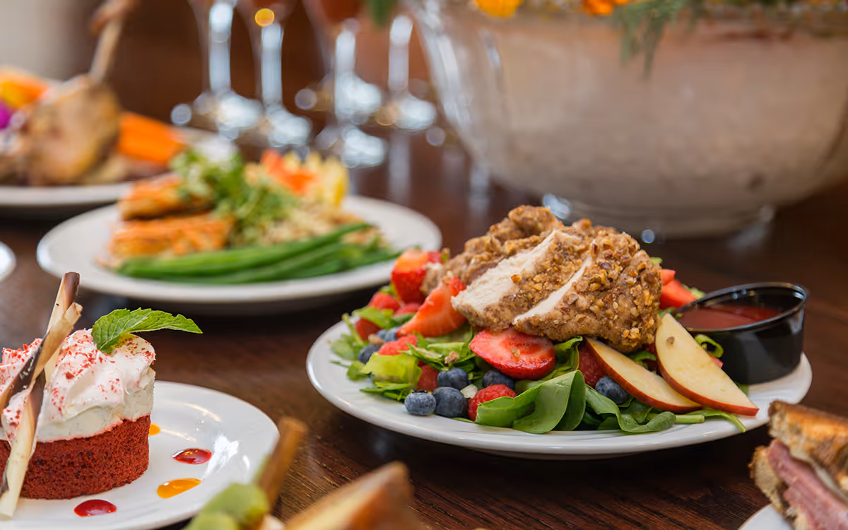 Close-up of a table with plates of food including a salad with sliced chicken, strawberries, blueberries, apple slices, and a small cup of dressing, a dessert with whipped cream and chocolate sticks, and other dishes in the background.