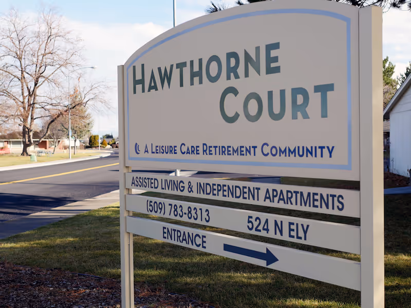 Outdoor sign for Hawthorne Court, a leisure care retirement community offering assisted living and independent apartments, located at 524 N Ely with a contact phone number (509) 783-8313. The sign is positioned near a road with some trees and buildings in the background.