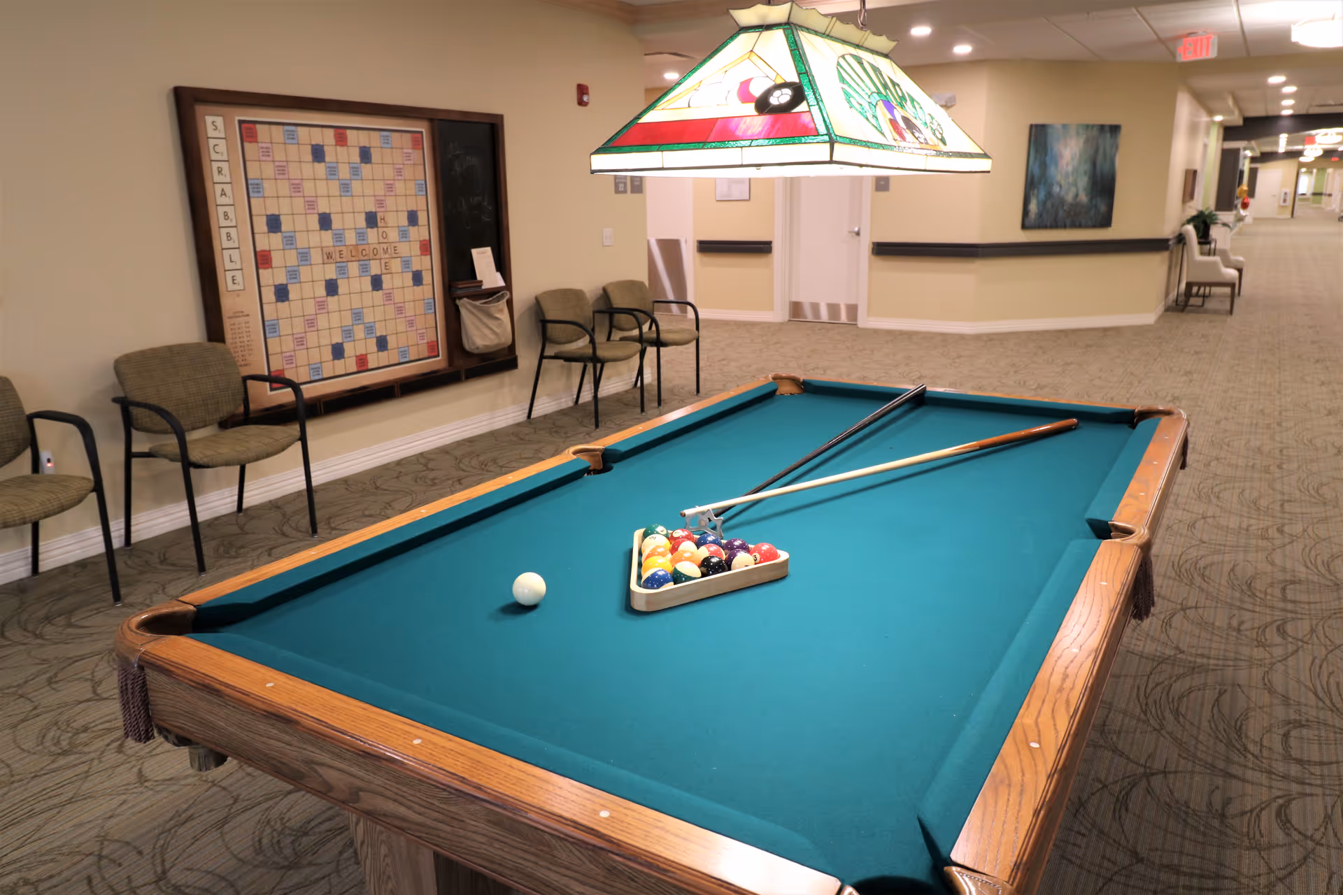 A pool table with racked balls and cues in a well-lit senior facility hallway with chairs and a Scrabble wall board.