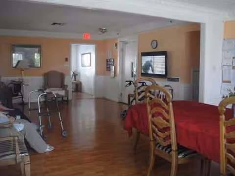 A shared senior living lounge with dining table, TV, chairs, and mobility aids.