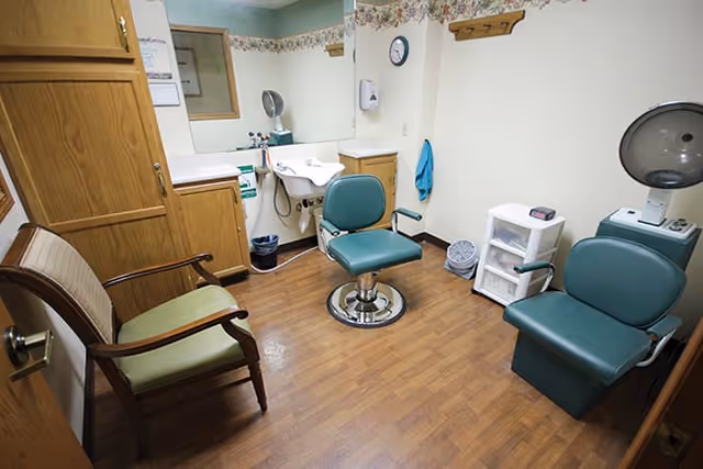 Interior of a small salon or grooming room with two green salon chairs, a wooden chair with green cushion, a sink with mirror, and a hair dryer hood. The room has wood flooring and light-colored walls with floral wallpaper border near the ceiling.