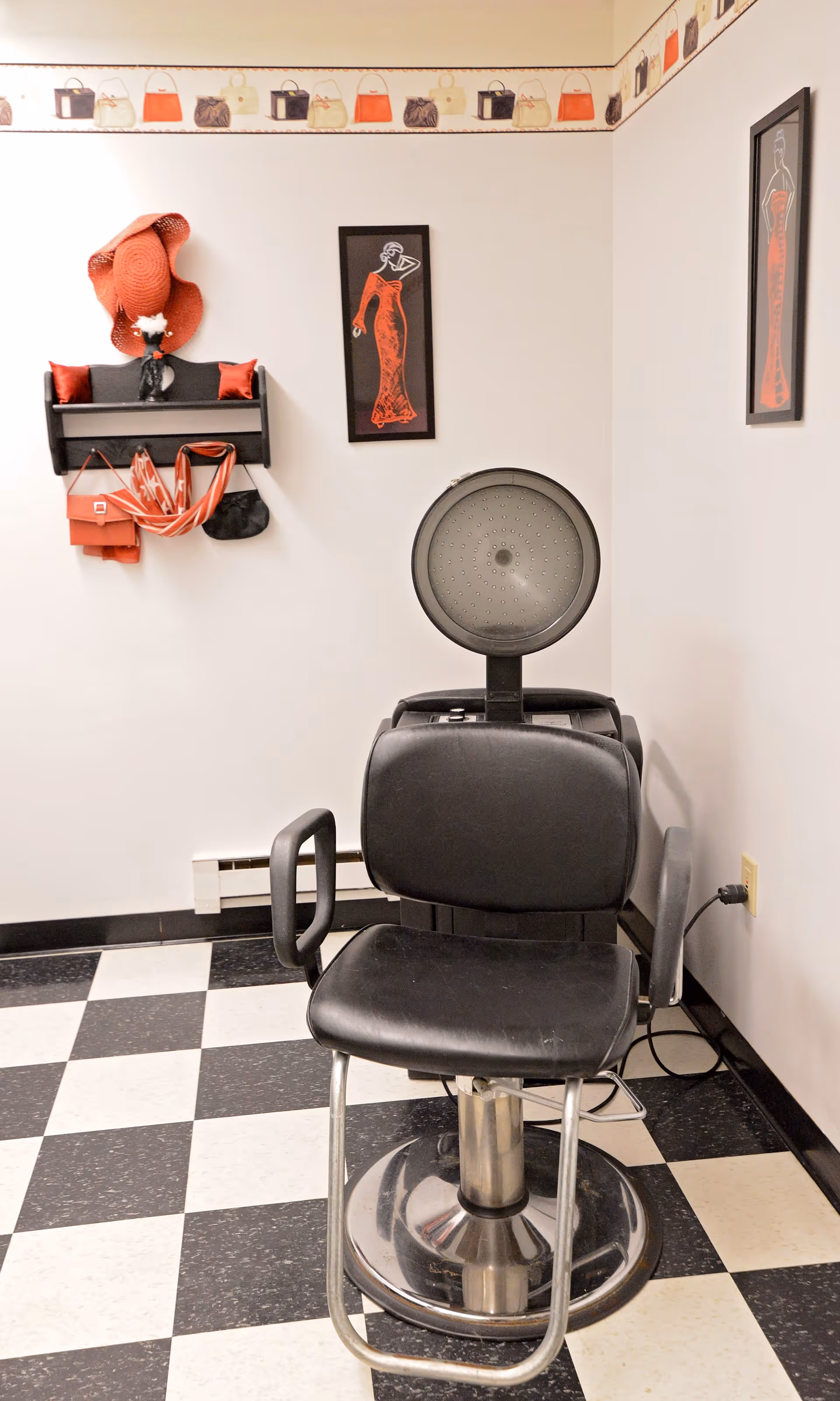 A black salon chair with a hooded hair dryer in a room with black and white checkered floor tiles. The walls are decorated with framed pictures of a woman in a red dress and a shelf holding red and black hats, purses, and scarves. There is a wallpaper border featuring various handbags near the ceiling.