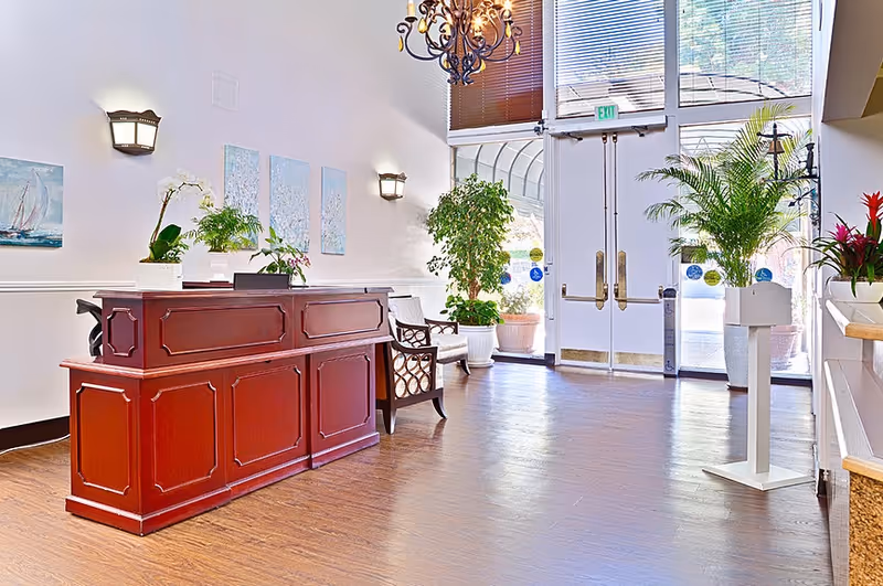 Bright lobby reception area with a wooden reception desk, seating, potted plants, chandelier, and glass double doors to the outside.