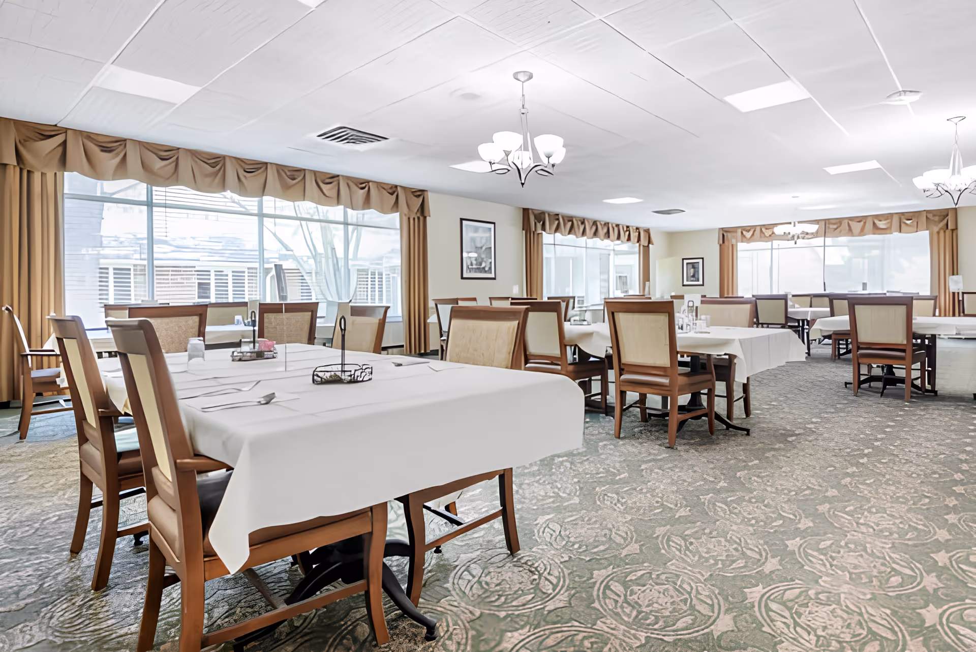 A spacious dining room with multiple tables covered in white tablecloths, each surrounded by wooden chairs with cushioned seats. Large windows with beige curtains allow natural light to fill the room. The carpet has a floral pattern, and chandeliers hang from the ceiling providing additional lighting.