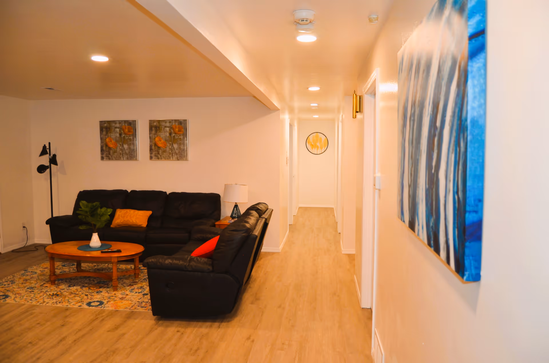 Interior view of a living room area with a black leather sectional sofa adorned with orange and red pillows, a wooden coffee table on a patterned rug, two framed floral paintings on the wall, a floor lamp, and a hallway leading to other rooms with additional wall art.