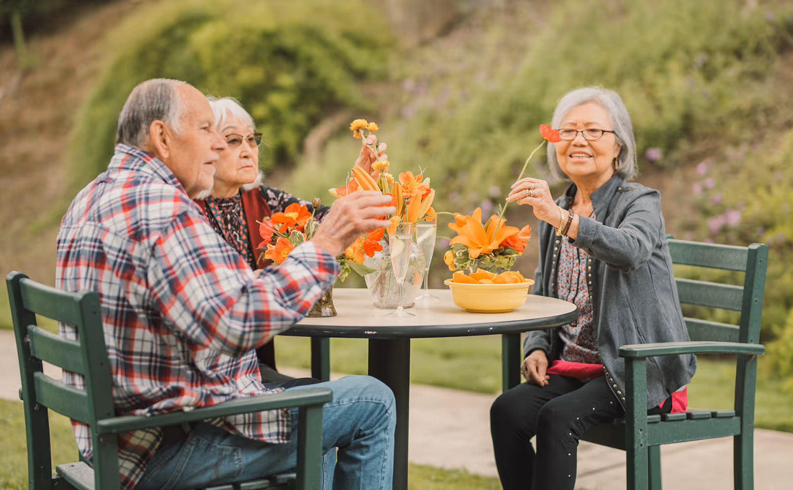 Three elderly people sitting around a round outdoor table with flower arrangements, smiling and holding flowers in a garden setting.