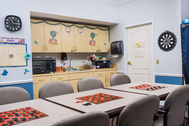 A room with a long table and several chairs arranged around it. On the table are three checkerboards with red and black checkers set up for play. The back wall has yellow cabinets decorated with pumpkin and wreath designs, a microwave, a toaster oven, and a paper towel dispenser. A dartboard is mounted on the wall next to a closed door. A clock and a July calendar are visible on the left wall.