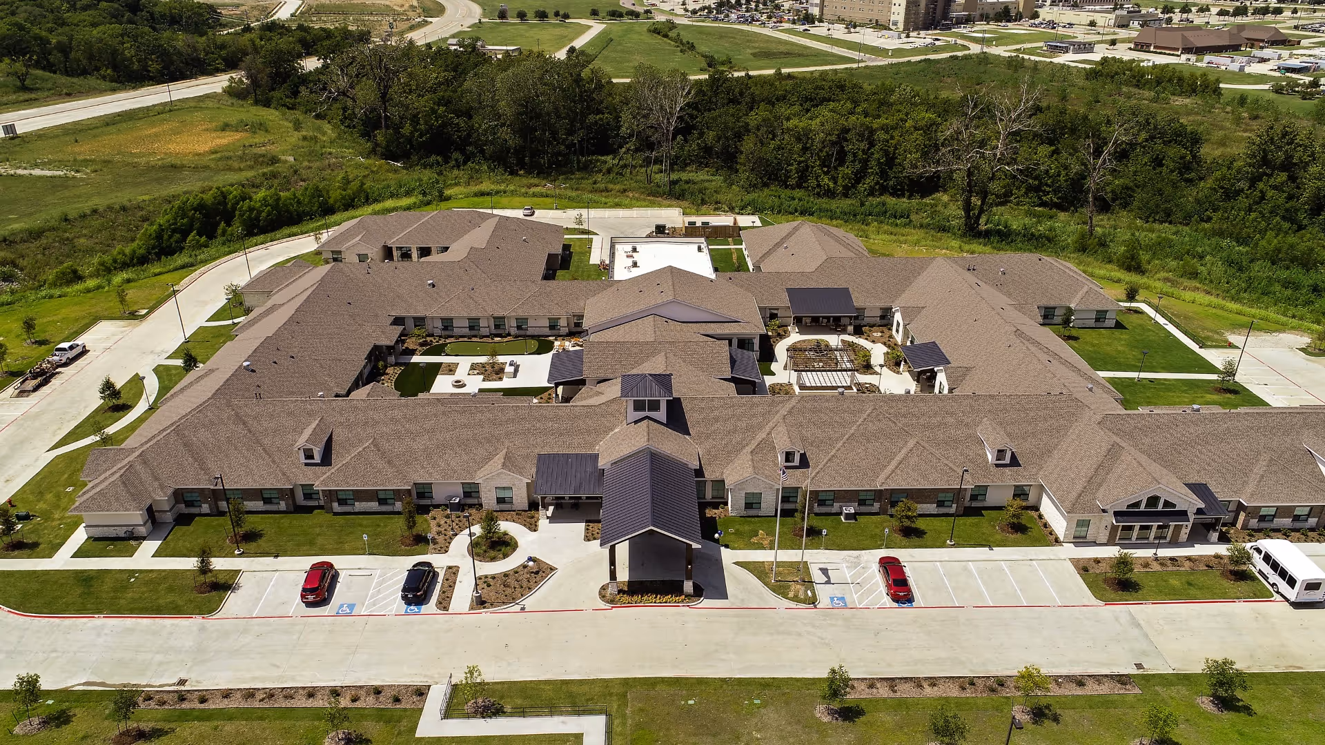Aerial view of Celebration Senior Living of Denison, showing a large, single-story building with multiple wings, surrounded by green lawns and trees. The building has a beige exterior with a brown roof and several covered entrances. There are parking spaces, including handicapped spots, in front of the building.