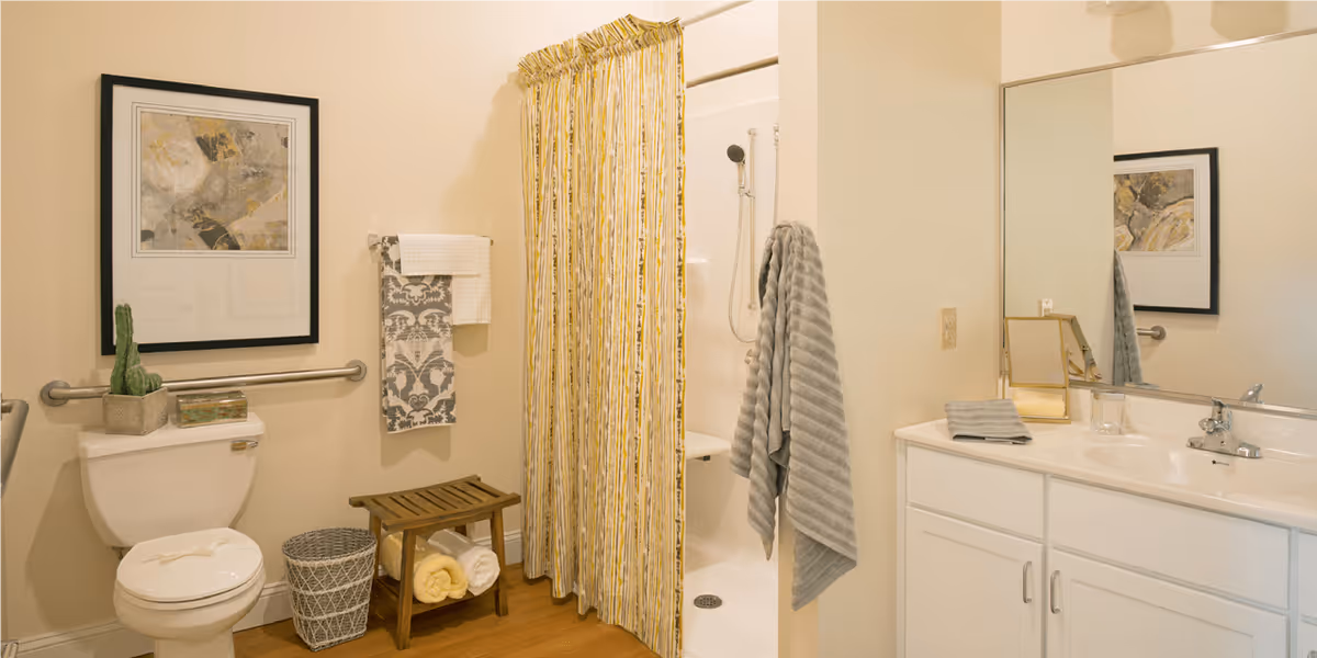 A clean and well-lit bathroom featuring a toilet with a small potted cactus and decorative box on the tank, a wall-mounted grab bar, a wooden bench with rolled towels underneath, a shower with a yellow and white striped curtain, a gray towel hanging on a hook, and a white vanity with a sink, mirror, and small accessories.