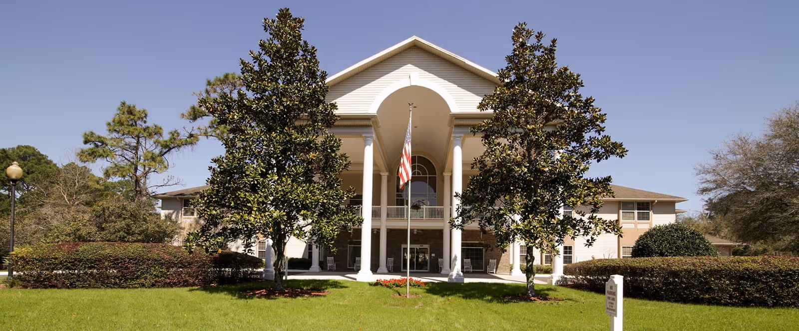 Front exterior view of a senior living facility building with white columns, an American flag on a flagpole in the center, surrounded by green grass, bushes, and trees under a clear blue sky.
