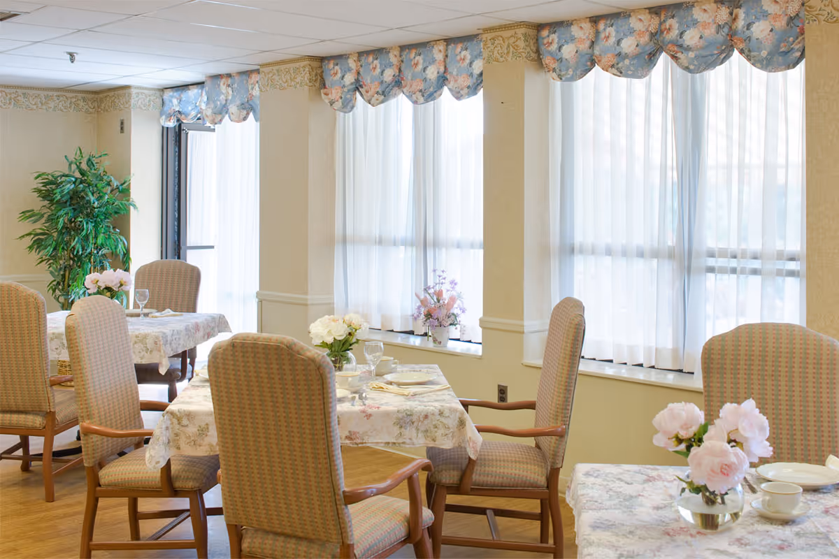 A bright dining room with several tables covered in floral tablecloths, each set with plates, cups, and glassware. Comfortable upholstered chairs surround the tables. Large windows with sheer white curtains and floral valances allow natural light to fill the room. A green potted plant and floral centerpieces add a touch of decoration.