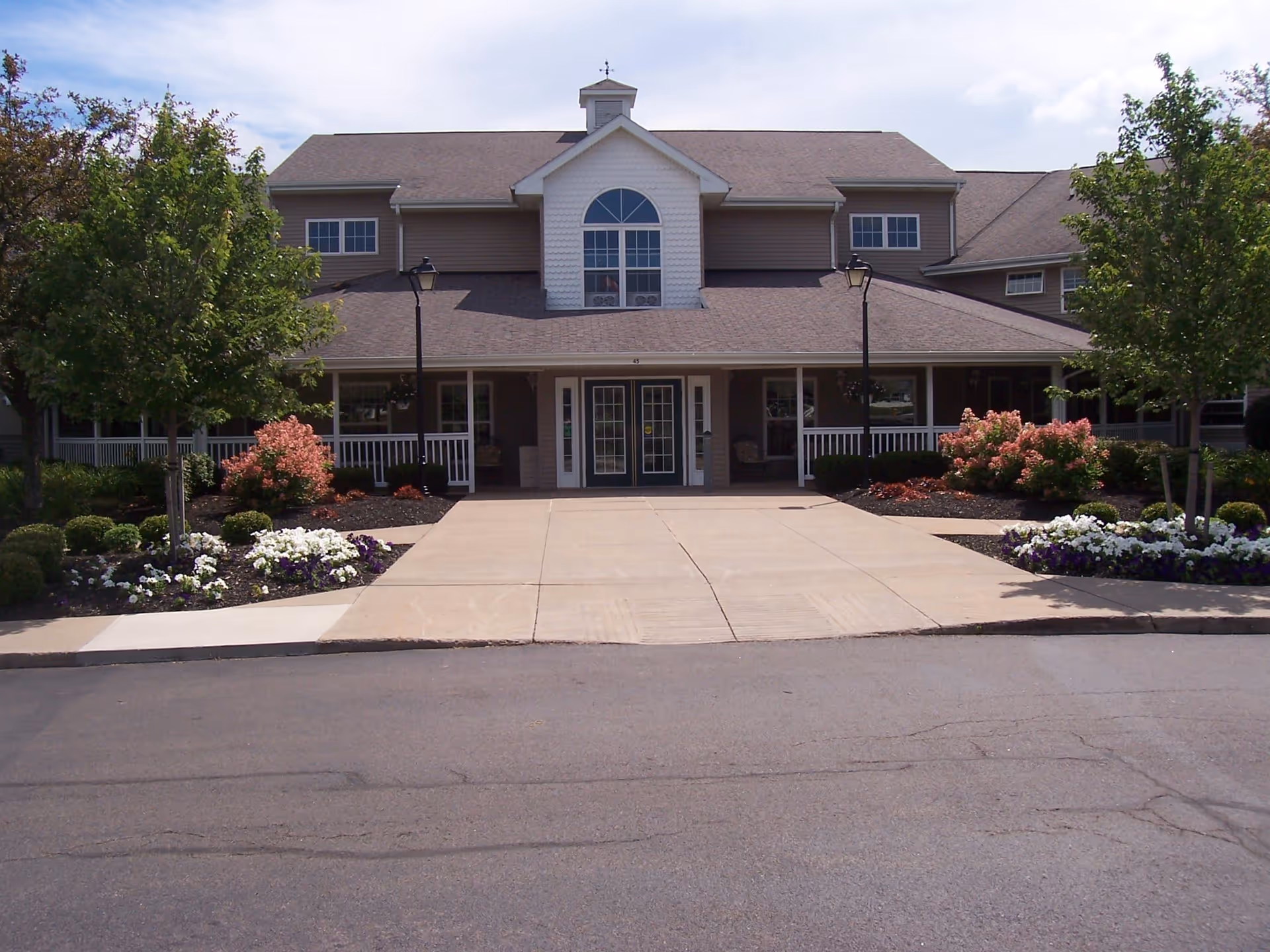 Front exterior view of College Greene Senior Apartments building with a paved driveway, landscaped flower beds with white and purple flowers, bushes, and trees on either side. The building has a large covered porch with white railings and multiple windows, under a gray shingled roof with a small cupola on top.