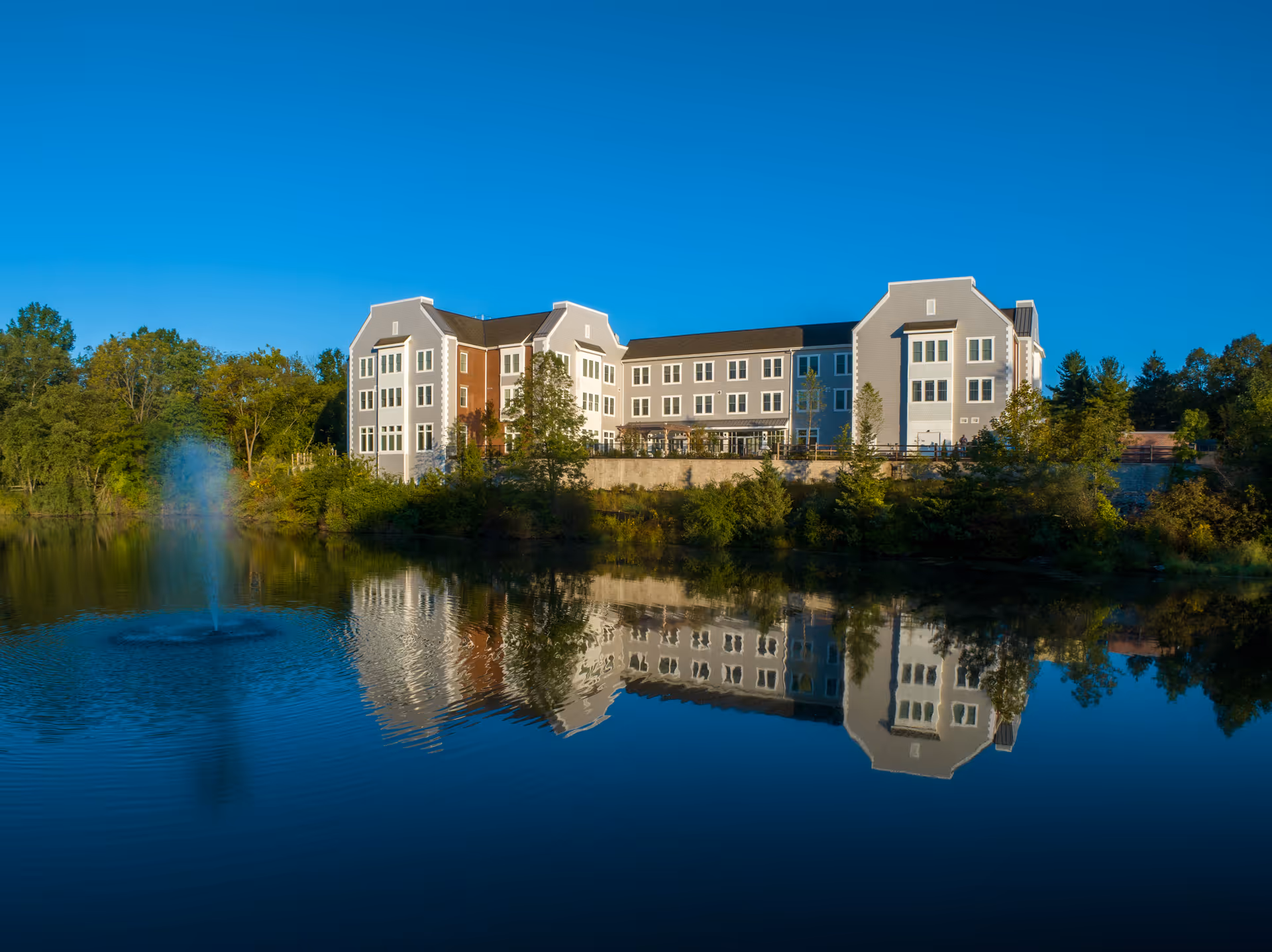 A multi-story residence building overlooks a pond with a fountain and its reflection in the water.