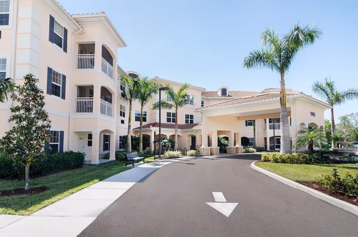 Exterior view of a senior living facility with a light peach-colored building, multiple windows, balconies, palm trees, a driveway with an arrow pointing forward, and a covered entrance area.
