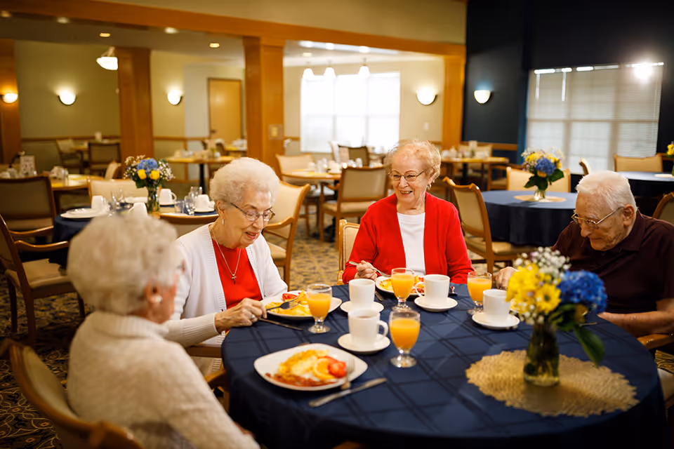 Four elderly residents sit around a table in a dining room enjoying breakfast with plates, coffee cups, and glasses of orange juice.