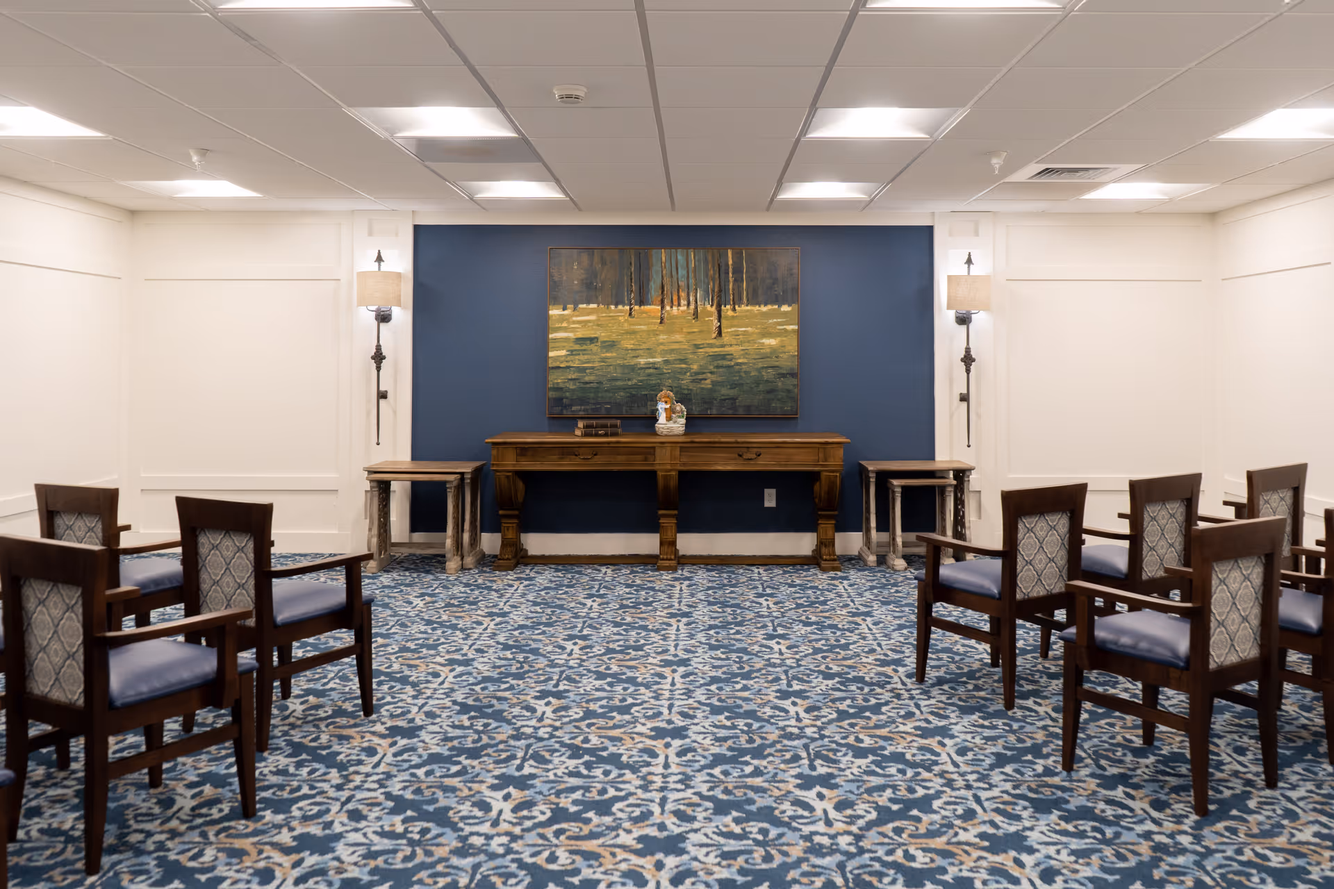 A small meeting or activity room with patterned blue carpet and white walls. There are eight wooden chairs with blue cushioned seats arranged in two rows facing a wooden console table against a blue accent wall. On the table are a few decorative items, and above it hangs a painting of a forest scene. Two wall-mounted lamps flank the table.