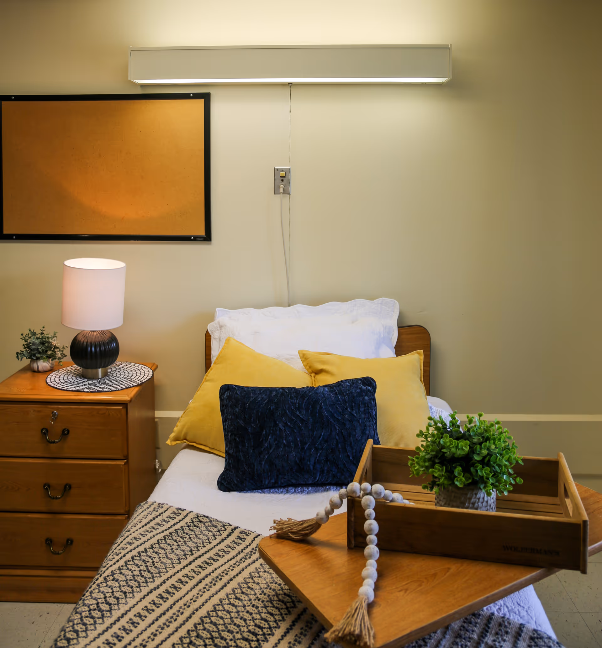 A neatly made single bed with white bedding, two yellow pillows, and one dark blue pillow. Next to the bed is a wooden nightstand with three drawers, a small lamp, and a small potted plant. On the bed is a wooden tray holding a small green plant and a decorative beaded garland. A cork bulletin board is mounted on the beige wall above the nightstand.