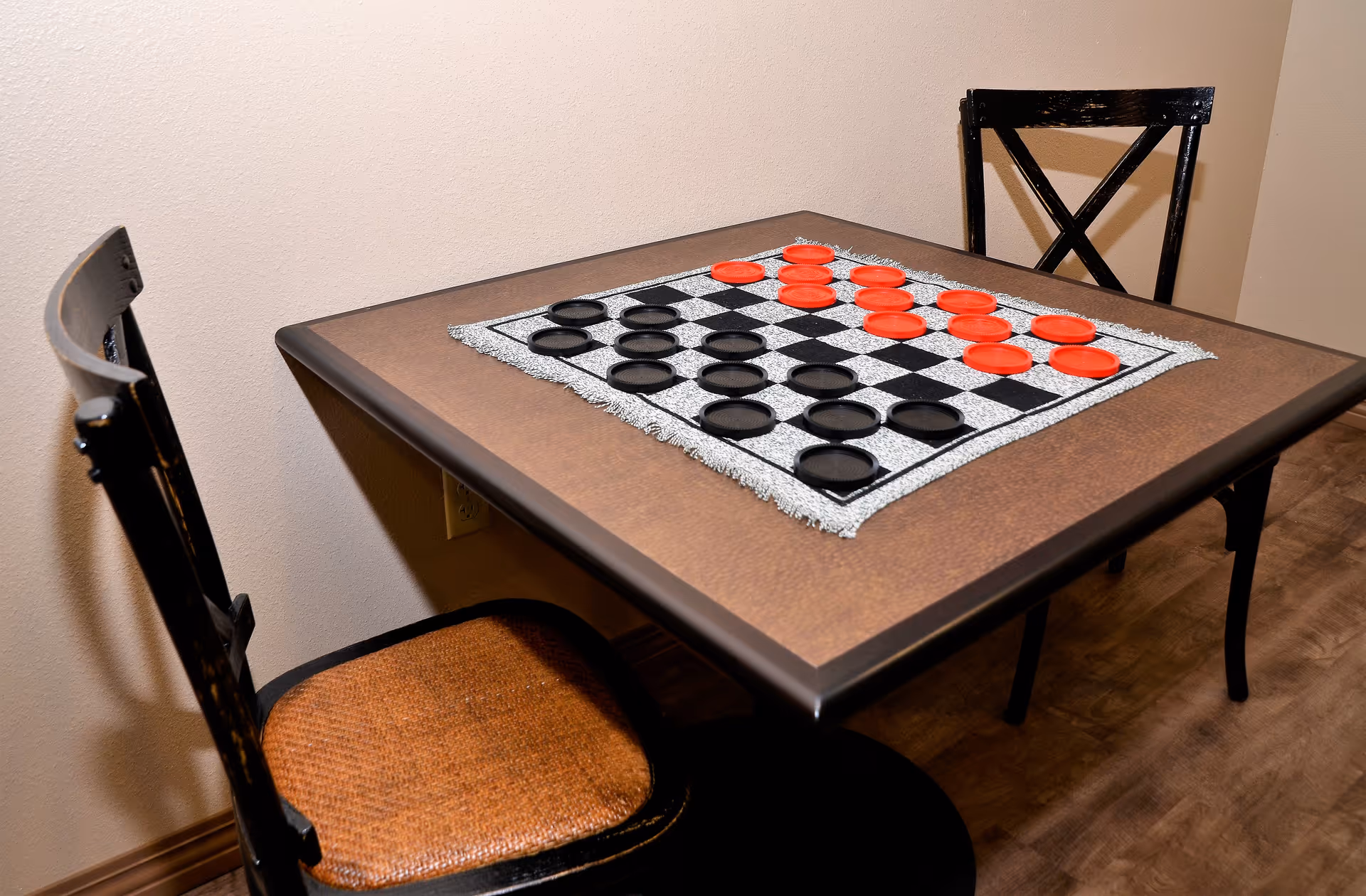 A small square table with a checkers board set up on a cloth, featuring red and black checkers pieces. Two wooden chairs with woven seats are positioned on opposite sides of the table. The setting is indoors with a plain beige wall and wood flooring.