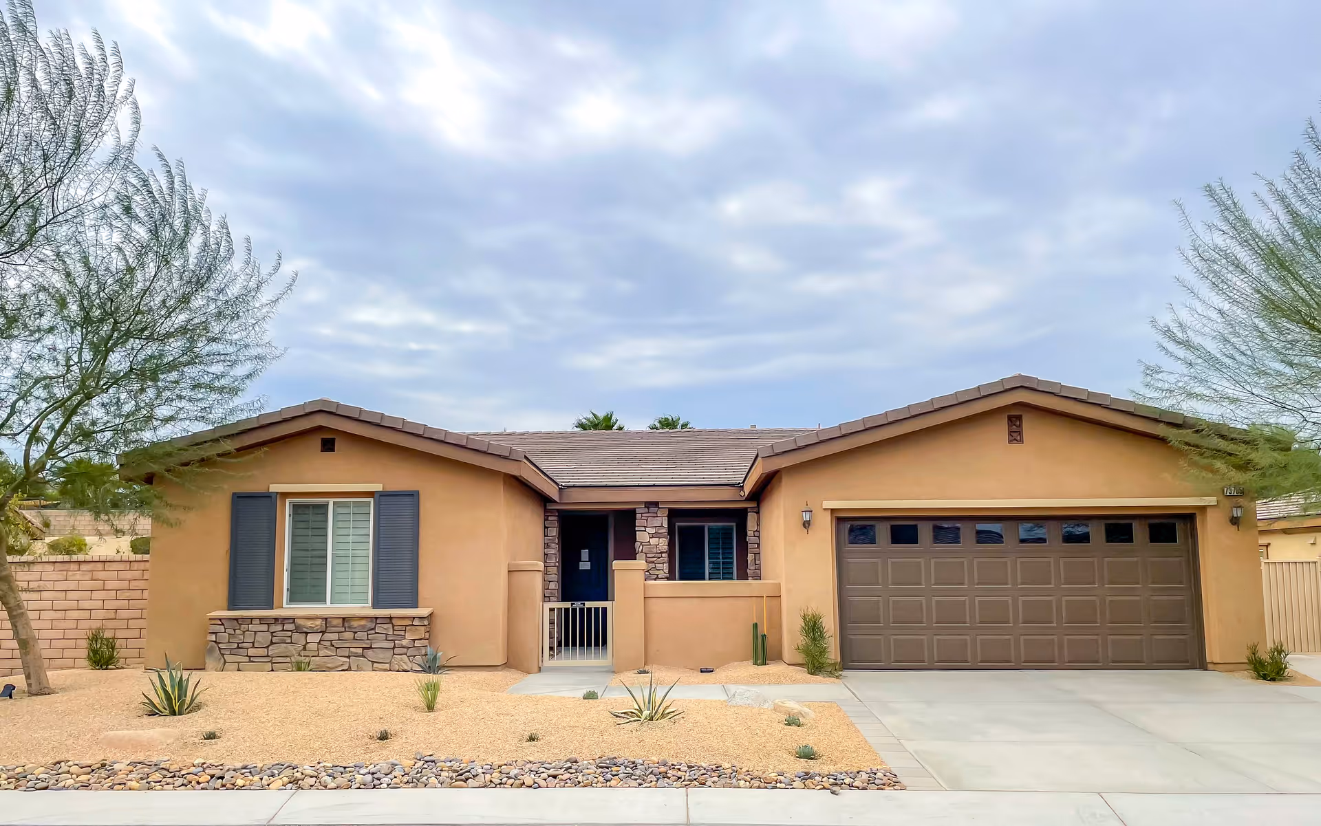 Single-story tan stucco house with an attached two-car garage and desert landscaping under a cloudy sky.