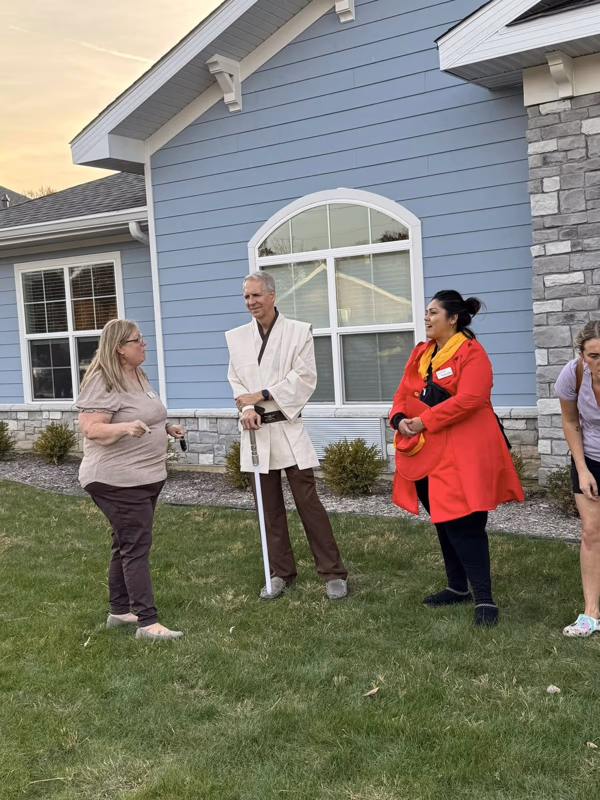 Three adults standing on a grassy lawn outside a blue building with stone accents. One man is dressed in a costume resembling a Jedi robe and holding a lightsaber prop. Two women are standing nearby, one wearing a red coat and the other in casual clothing. They appear to be engaged in conversation.