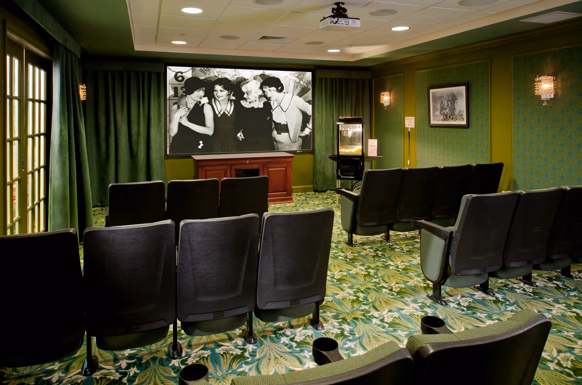 A small movie theater room with rows of black cushioned seats facing a large screen displaying a black and white image of four women. The room has green patterned carpet and green walls with sconces. There is a popcorn machine on the right side near the screen and a wooden cabinet below the screen.