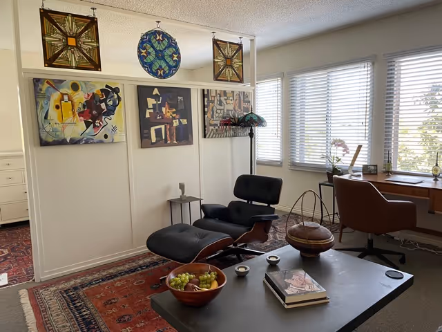 A cozy living room area with a black lounge chair and ottoman, a wooden desk with a brown chair near windows with blinds, and a coffee table holding a bowl of fruit, two small cups, and a book. The walls are decorated with colorful abstract paintings and a stained glass piece hanging from the ceiling. A patterned rug covers part of the floor.